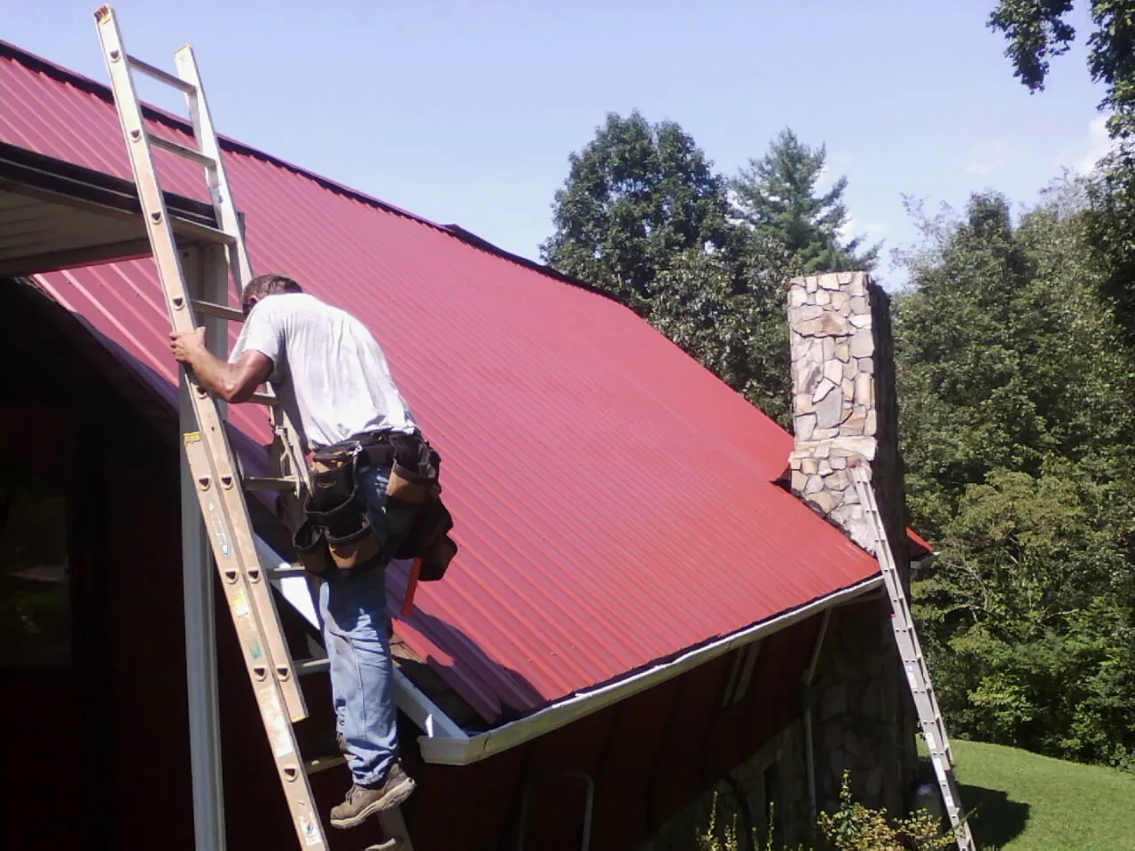 Roofer on a ladder beside a red metal roof and stone chimney — Soffit & Fascia Repair by Roofing Brothers in Little Hocking